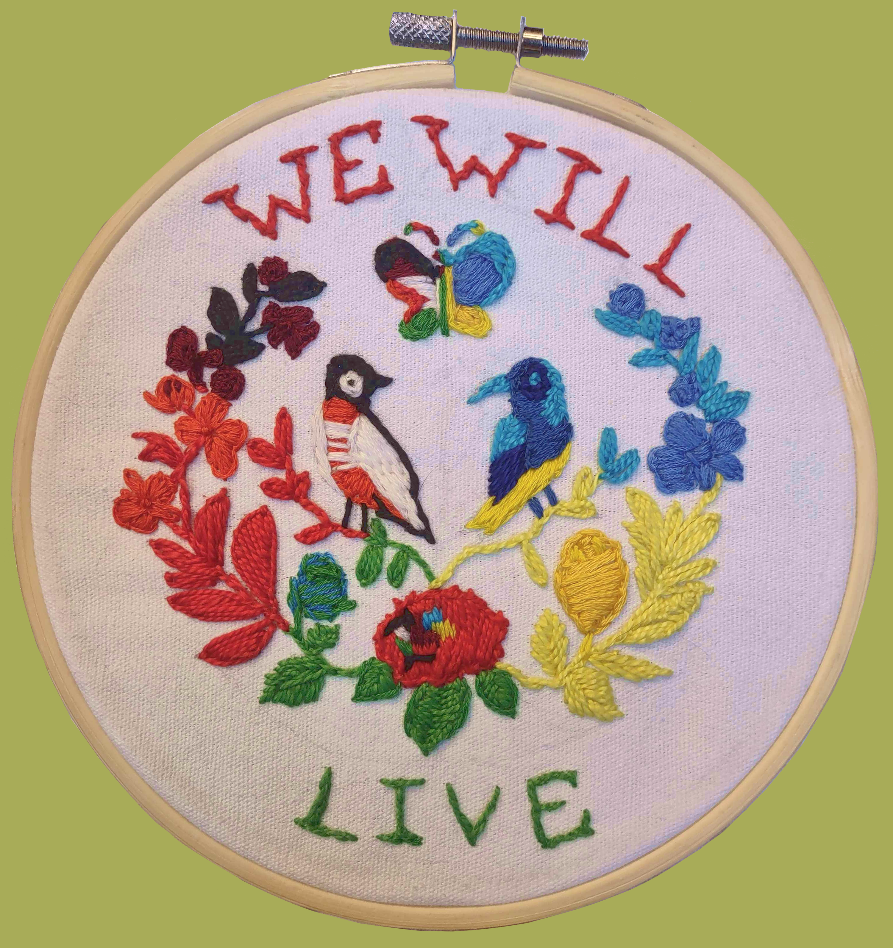 Two embroidered birds on flowers and leaves with a butterfly in the center, saying'We will live' around the edges.